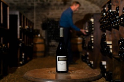 A bottle of Premier Cru Givry wine sitting on a table in front of a man reaching for a bottle of wine in a wine cellar.